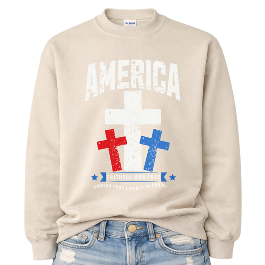 Christian patriotic faith graphic on a beige sweatshirt. The design features a bold distressed “America” arch across the chest with a large central white cross, flanked by red and blue crosses below for a strong USA color story. A banner reading “Faithful and Free” anchors the design, with smaller text noting “Finding True Liberty in Christ,” blending faith-forward messaging with classic Fourth of July patriotic style.