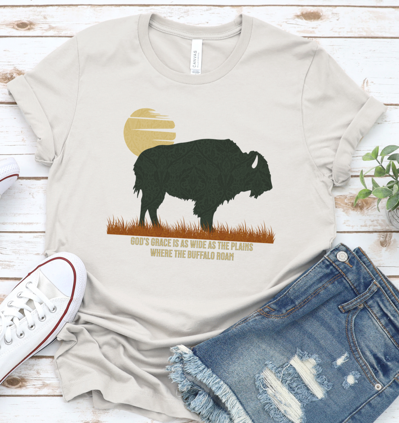 A beige unisex graphic t-shirt with a buffalo silhouette design, accompanied by text, laid out flat with a pair of blue denim jeans and a white sneaker. God's Grace is as wide as the plains where the buffalo roam.