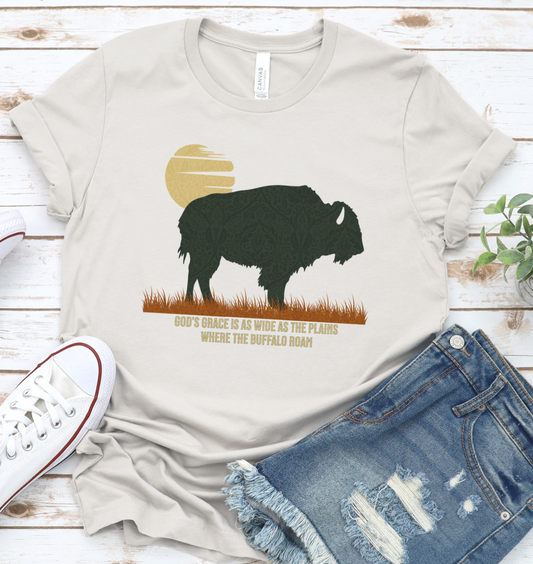 A beige unisex graphic t-shirt with a buffalo silhouette design, accompanied by text, laid out flat with a pair of blue denim jeans and a white sneaker. God's Grace is as wide as the plains where the buffalo roam.