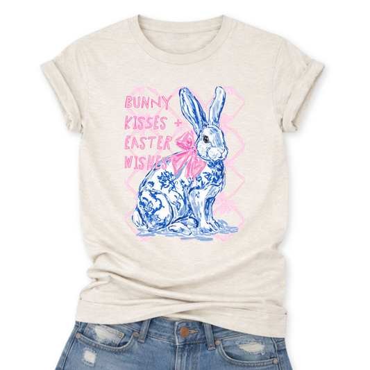 Easter spring graphic on a Bella Canvas t-shirt. The design features a hand-illustrated bunny in blue toile-style floral detailing, accented with a soft pink bow and layered over a subtle bow-pattern background. The phrase “Bunny Kisses + Easter Wishes” appears in playful pastel lettering, blending classic spring charm with boutique-ready styling.