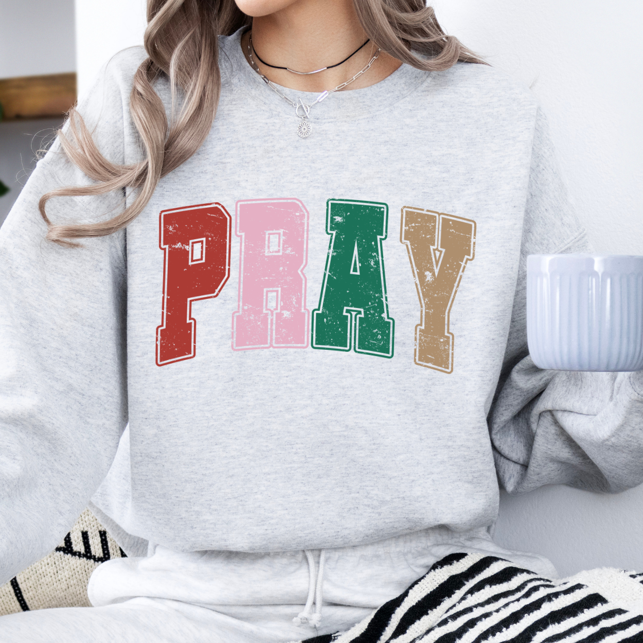 Person wearing a Ash gray sweatshirt with the words PRAY across the chest with the colors red, pink, green, tan