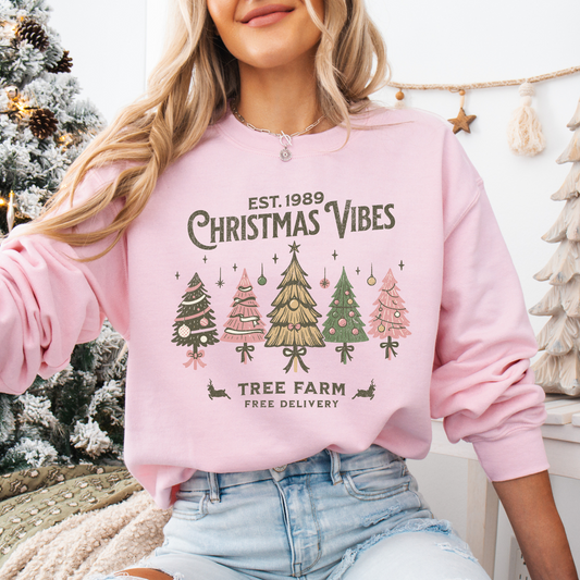 Person wearing a pink sweatshirt with Christmas tree graphics and text in a festive setting.  Shirt says Est 1989 Christmas vibes tree farm free delivery