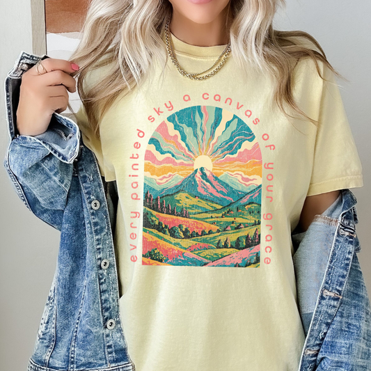 Person wearing a yellow t-shirt with a colorful mountain graphic and text.  Every painted sky a canvas of your grace on the shirt with a beautiful mountain sunset pink and blue graphics on a yellow t-shirt