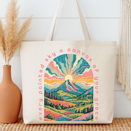 Tote bag with mountain design and text next to a vase on a wooden surface.  Every painted sky a canvas of your grace