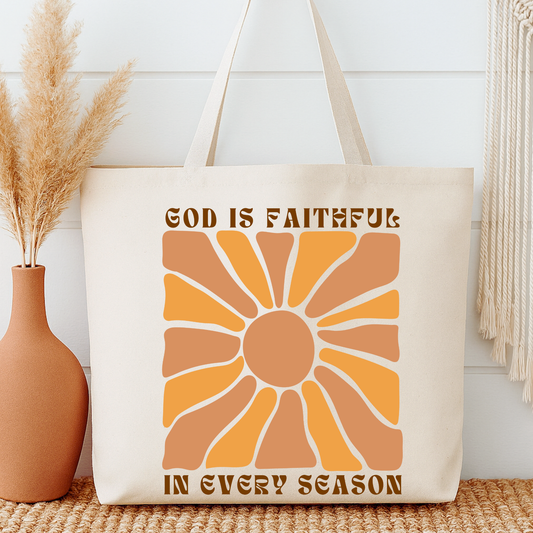 Tote bag with sun design and text 'God is faithful in every season' next to a vase with dried plants.