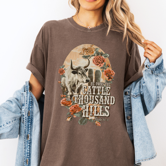 Person wearing a brown t-shirt with a graphic design and text, holding a denim jacket.  He owns the cattle on a thousand hills Psalm 50:10