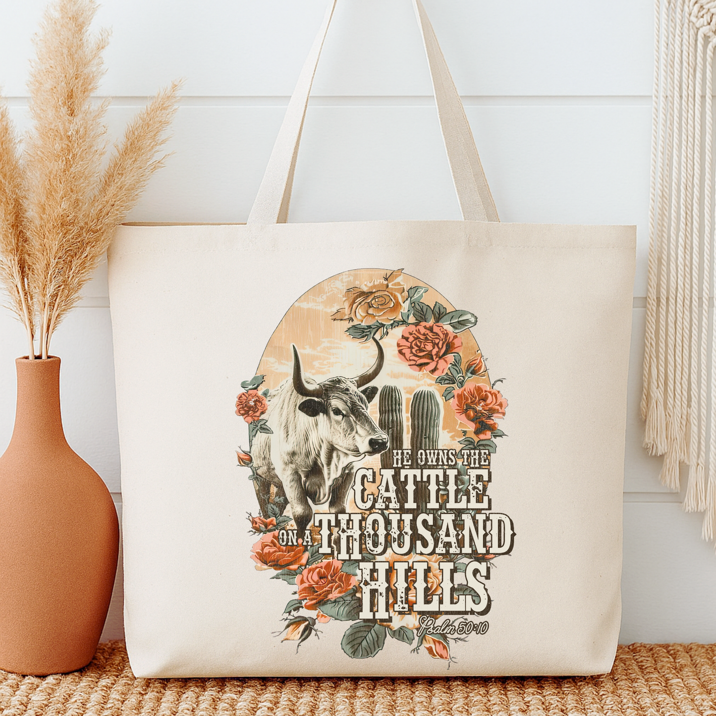 Tote bag with a bull and floral design, featuring text, next to a vase with dried plants.  He owns the cattle on a thousand hills.