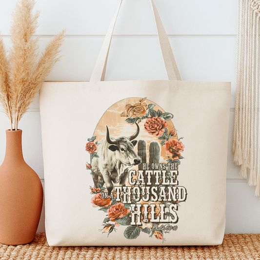 Tote bag with a bull and floral design, featuring text, next to a vase with dried plants.  He owns the cattle on a thousand hills.
