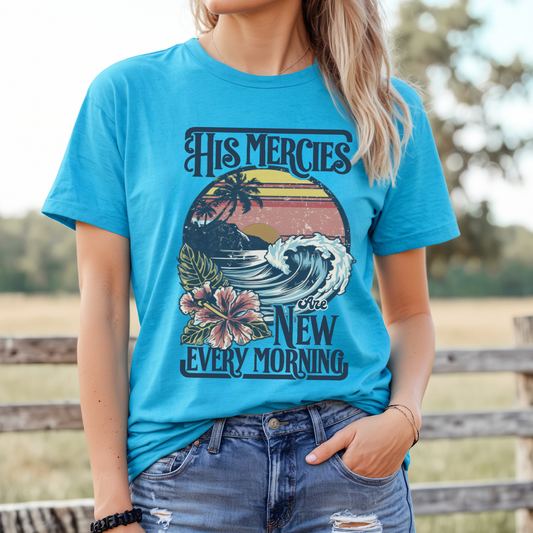 A person wearing a blue graphic t-shirt with text and a tropical design, including waves and flowers, standing in front of a wooden fence.  Graphic says His Mercies are new every morning.