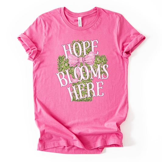 Christian spring cross graphic on a Bella Canvas t-shirt. The design features a greenery-filled cross adorned with a large pink bow, layered with the phrase “Hope Blooms Here” in bold, vintage-inspired lettering. The artwork blends Easter symbolism with soft feminine styling, creating a faith-forward seasonal piece. 