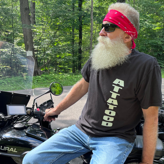 An older guy with a white beard and red bandana across his head sitting on a motorcyle wearing a black t-shirt that says  ATTA TOOD vertical up and down the shirt.