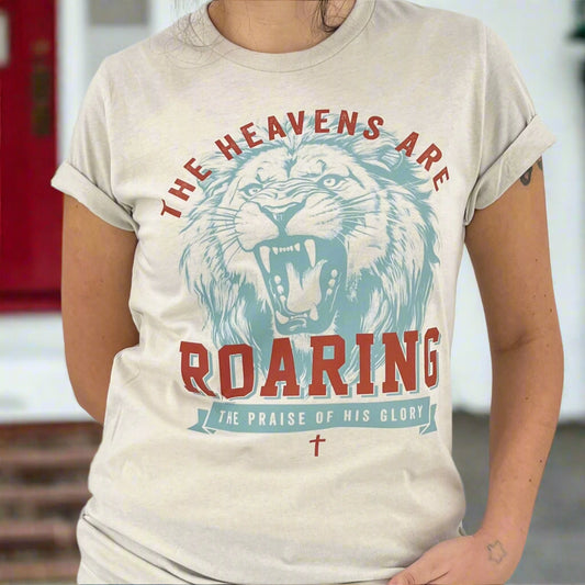 Christian t-shirt that says The heavens are roaring.  The praise of his glory.  Blue large lion with red lettering on a cream color Bella Canvas t-shirt