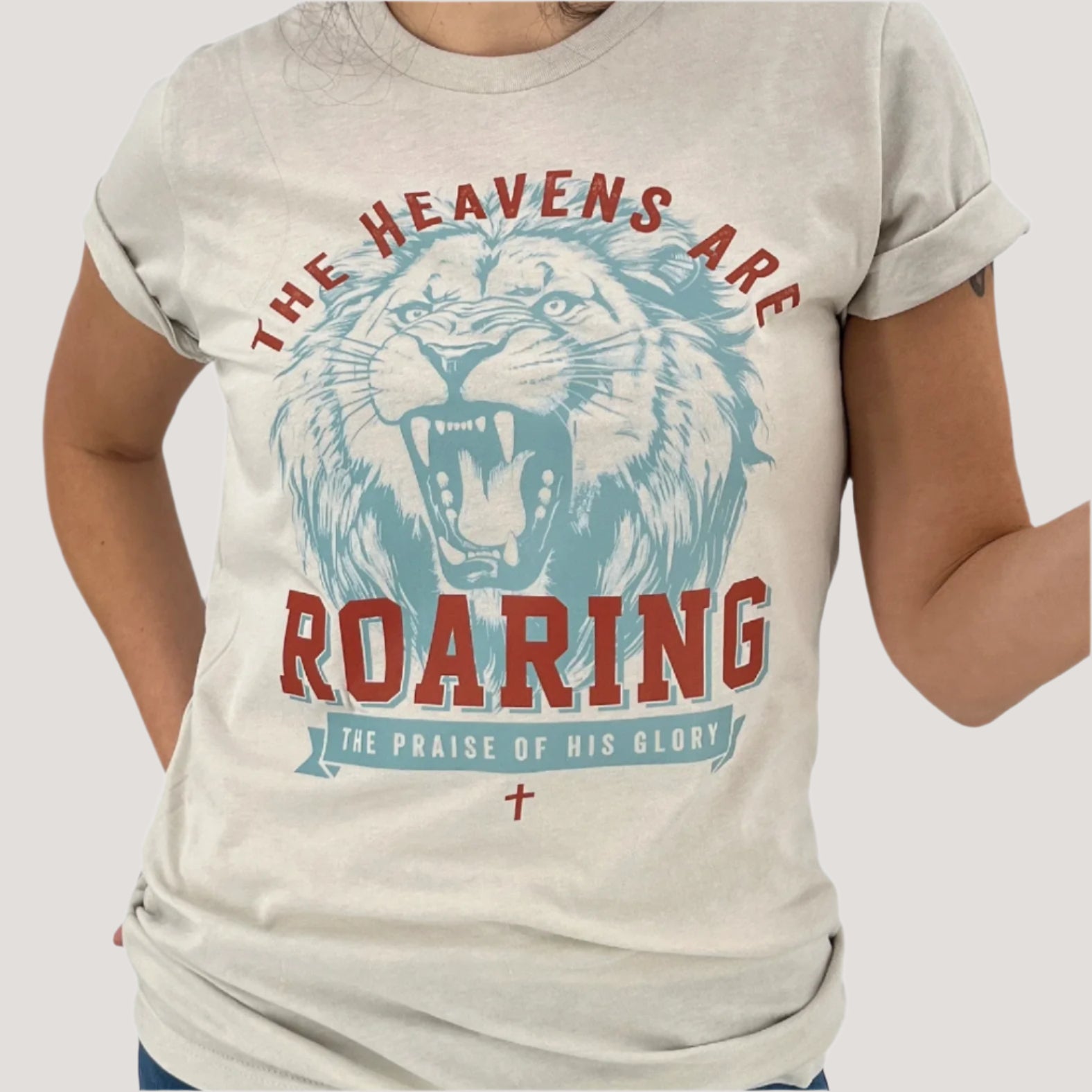Christian t-shirt that says The heavens are roaring.  The praise of his glory.  Blue large lion with red lettering on a cream color Bella Canvas t-shirt