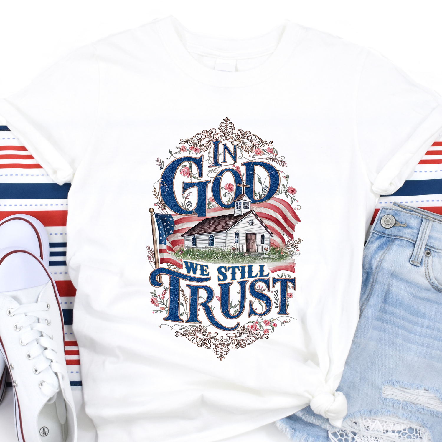 White t-shirt with 'In God We Still Trust' design featuring an American flag and small building, displayed with shoes and jeans.