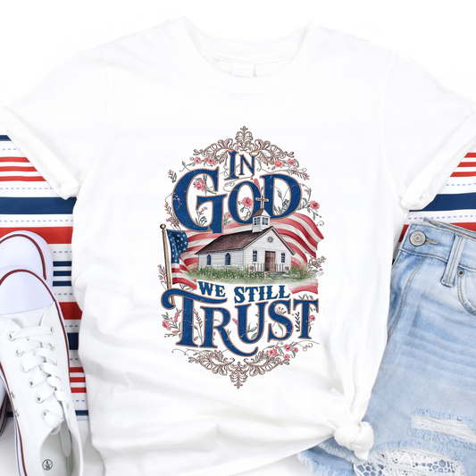 White t-shirt with 'In God We Still Trust' design featuring an American flag and small building, displayed with shoes and jeans.