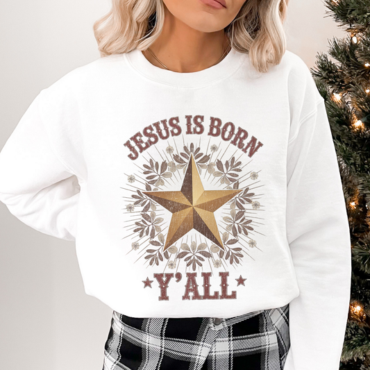 White sweatshirt with 'Jesus is Born Y'all' design worn by a person, Christmas tree in the background