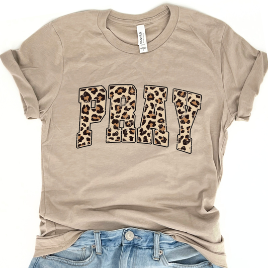 A beige PRAY graphic t-shirt with leopard print text on it, displayed on a flat surface with casual clothing.
