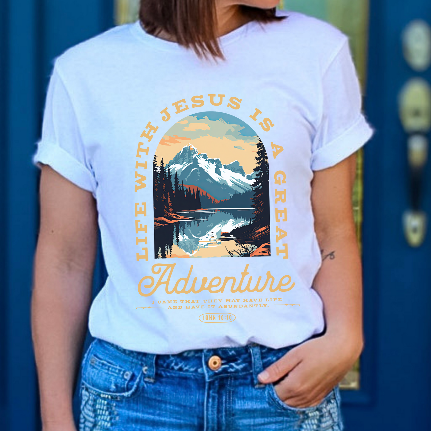A person wearing a white graphic t-shirt with text and a mountainous landscape design. Scripture text Life with Jesus is a Great Adventure John 10:10