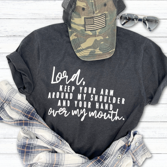 A graphic t-shirt with the text 'Lord, keep your arm around my shoulder your hand over my mouth' printed on the front, laid out flat with a camouflage hat, a pair of sunglasses, and a plaid shirt.
