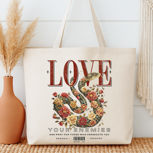 Tote bag with floral snake design and text next to a vase on a wooden surface.  Lover your enemies and pray for those who persecute you.