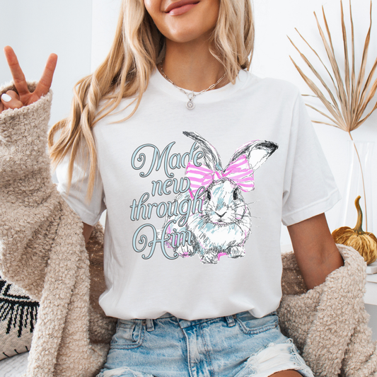 Christian Easter resurrection graphic on a Bella Canvas t-shirt. The design features a hand-sketched bunny accented with a pink bow and soft pastel shading, paired with the phrase “Made New Through Him” in flowing, feminine lettering. The artwork highlights the renewal message of Christ and the hope of Easter, blending soft spring styling with a strong faith-forward statement.
