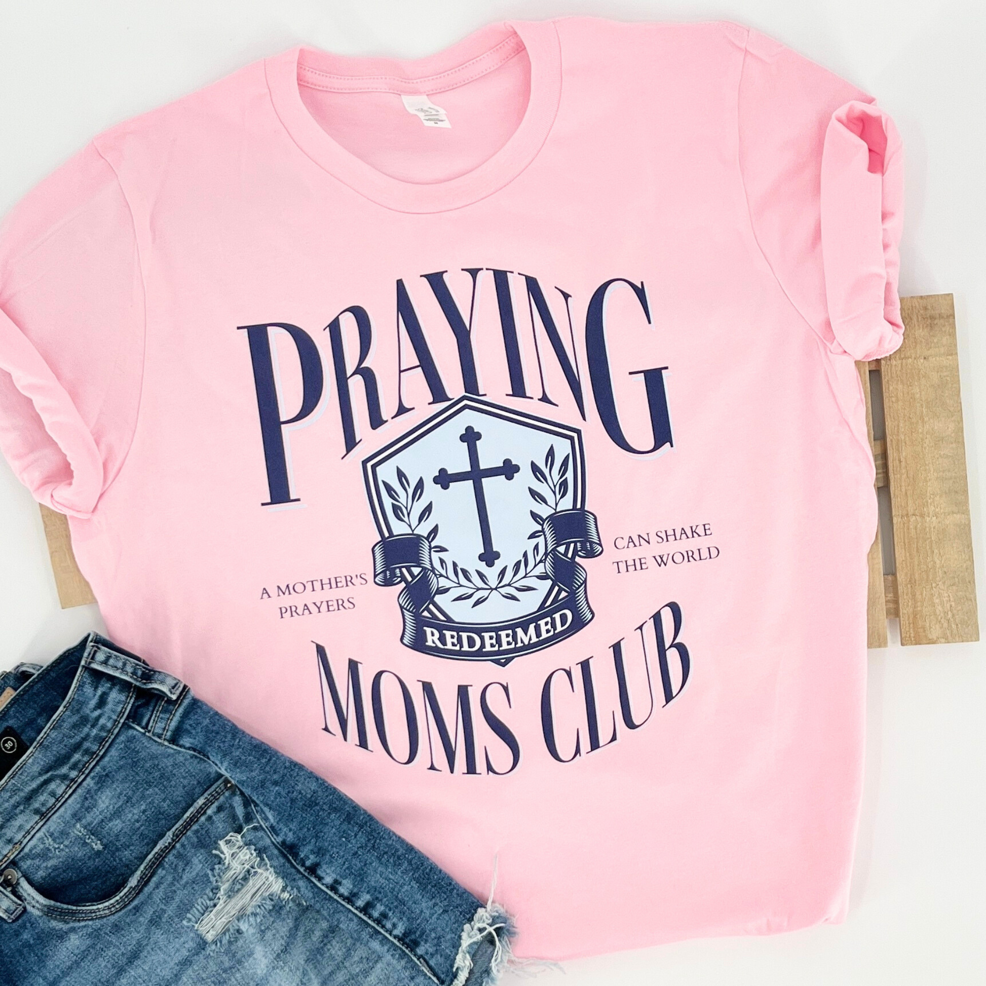 A pink graphic t-shirt with the text 'PRAYING MOMS CLUB' and a emblem on the front, displayed flat with a pair of blue denim jeans.