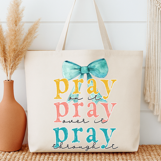 Beige tote bag with colorful text and bow design, placed on a wooden surface with decorative elements.  Pray on it pray over it pray through it