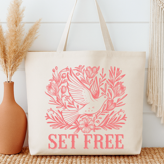 Beige tote bag with pink bird and floral design and 'SET FREE' text, next to a vase with dried plants.