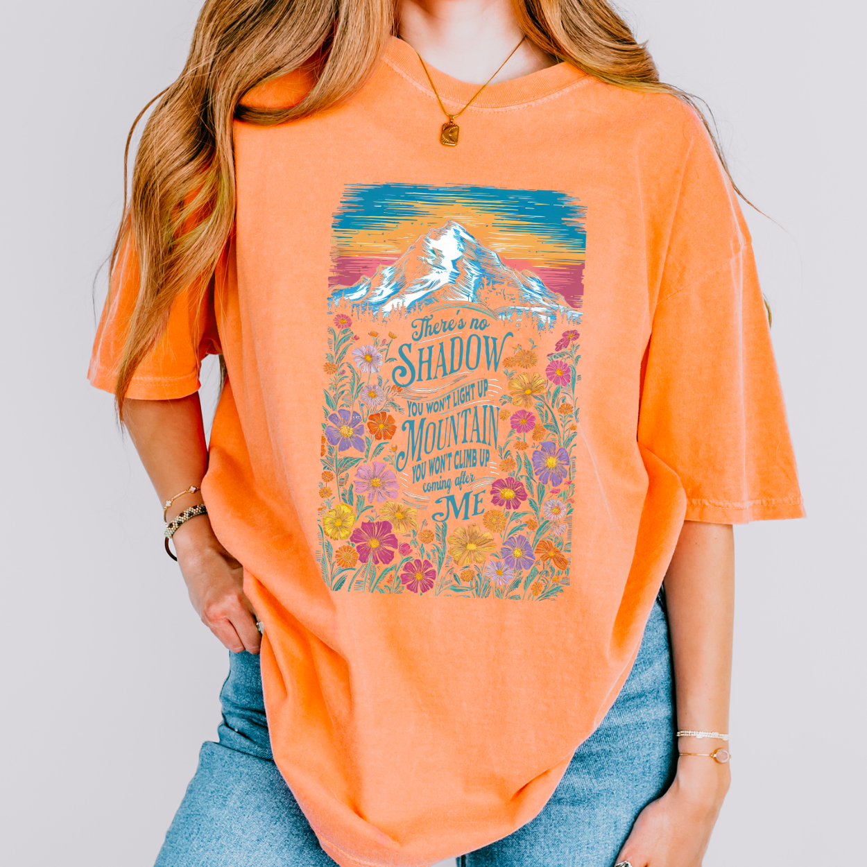 Person wearing an orange t-shirt with a mountain and floral design on a plain background.  There's no shadow you won't light up, Mountain you won't climb up coming after me