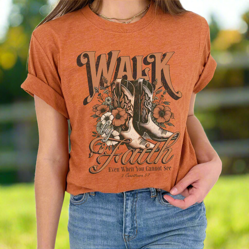 A person wearing a brown graphic t-shirt with text and a design that includes boots and flowers.  Text says Walk by Faith Even when you cannot see