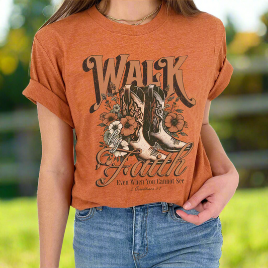 A person wearing a brown graphic t-shirt with text and a design that includes boots and flowers.  Text says Walk by Faith Even when you cannot see