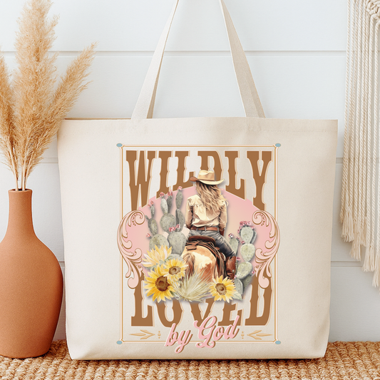 Tote bag with a design of a woman on a horse, cacti, and flowers with text 'Wildly Loved by God' next to a vase with dried plants.
