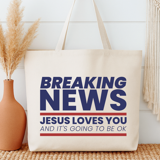 Beige tote bag with 'Breaking News: Jesus Loves You and It's Going to Be Ok' text, next to a vase with dried plants.
