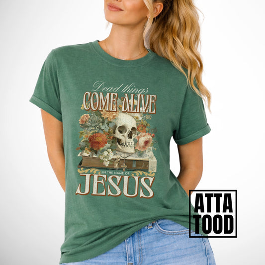 Dead things come alive in the name of Jesus with an oversized graphic of a bible, skeleton head and flowers on a comfort colors green tee