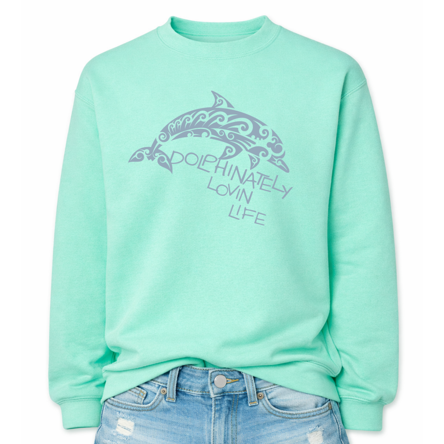 Coastal ocean graphic on a classic ivory mint green crewneck sweatshirt. The design features a large dolphin illustration filled with decorative swirl and wave inspired pattern details that give the sea life artwork a textured ocean aesthetic. The phrase Dolphinately Lovin Life is integrated into the graphic beneath the dolphin.