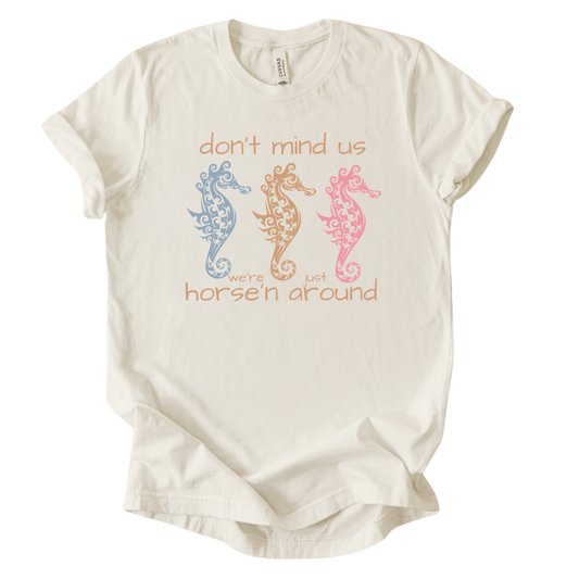 Coastal sea life graphic on a soft unisex heather oatmeal graphic tee. The design features three decorative seahorse illustrations arranged across the center of the artwork in soft coastal colors including blue, coral, and peach. Each seahorse contains detailed swirl and ocean pattern elements that give the sea life illustration a textured coastal aesthetic.