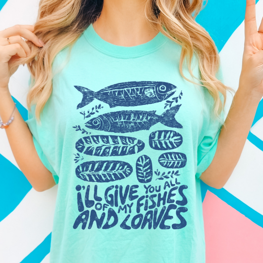 Person wearing a light blue t-shirt with fish illustrations and text against a colorful geometric background.  I'll give you all of my fishes and loaves.  Graphic has fish and loaves of bread