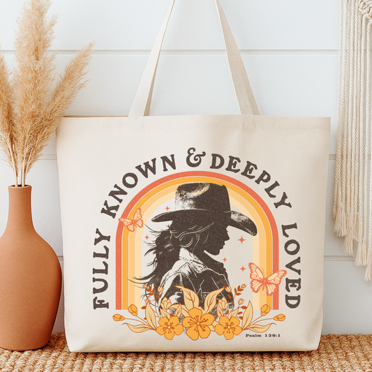Tote bag with a design of a woman in a hat, flowers, and butterflies with text 'Fully Known & Deeply Loved' on a neutral background.