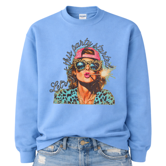 Patriotic Fourth of July graphic on a blue sweatshirt. The design features an illustrated woman wearing leopard-print sunglasses and a backward cap, holding a lit sparkler in her mouth. The artwork is layered with bold color contrasts, distressed textures, and playful script lettering that reads “Let’s Get This Party Started.” A high-energy patriotic design blending Americana attitude with modern pop-art style