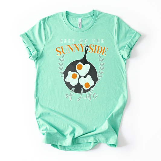 Bella Canvas unisex graphic t-shirt mockup with “Keep On The Sunny Side of Life” text and a sunny side up eggs design in a black skillet, with small leafy accents; bright, cheerful, retro-style positive quote tee.