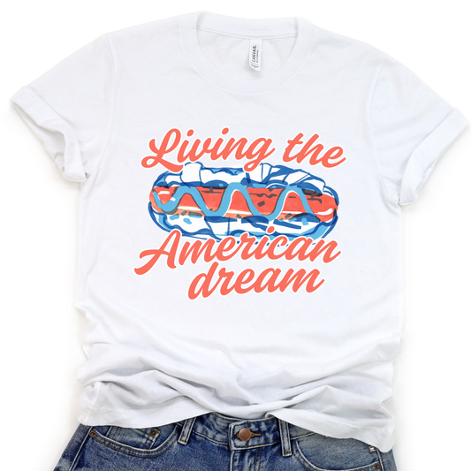 A white graphic t-shirt with a text 'Living the American dream' and a patterned design inside the text.  With a Red white and blue hot dog