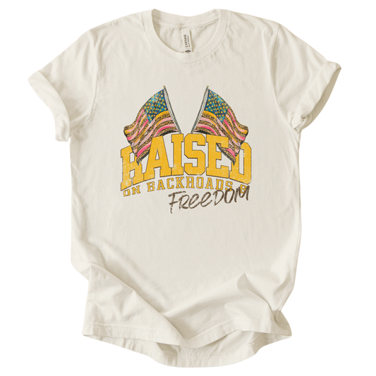 Patriotic Americana graphic on a t-shirt. The design features bold varsity-style “RAISED” lettering with crossed American flags above, finished with “on backroads” and “Freedom” in script below. The distressed, vintage-inspired texture and warm tones make it an easy Fourth of July t-shirt. Cream color tee