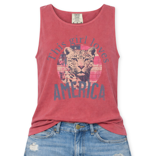 Crimson Red Comfort Colors tank top featuring a vintage-style patriotic graphic with a leopard illustration inside a distressed American flag circle and the phrase “This Girl Loves America,” styled with light wash denim jeans.