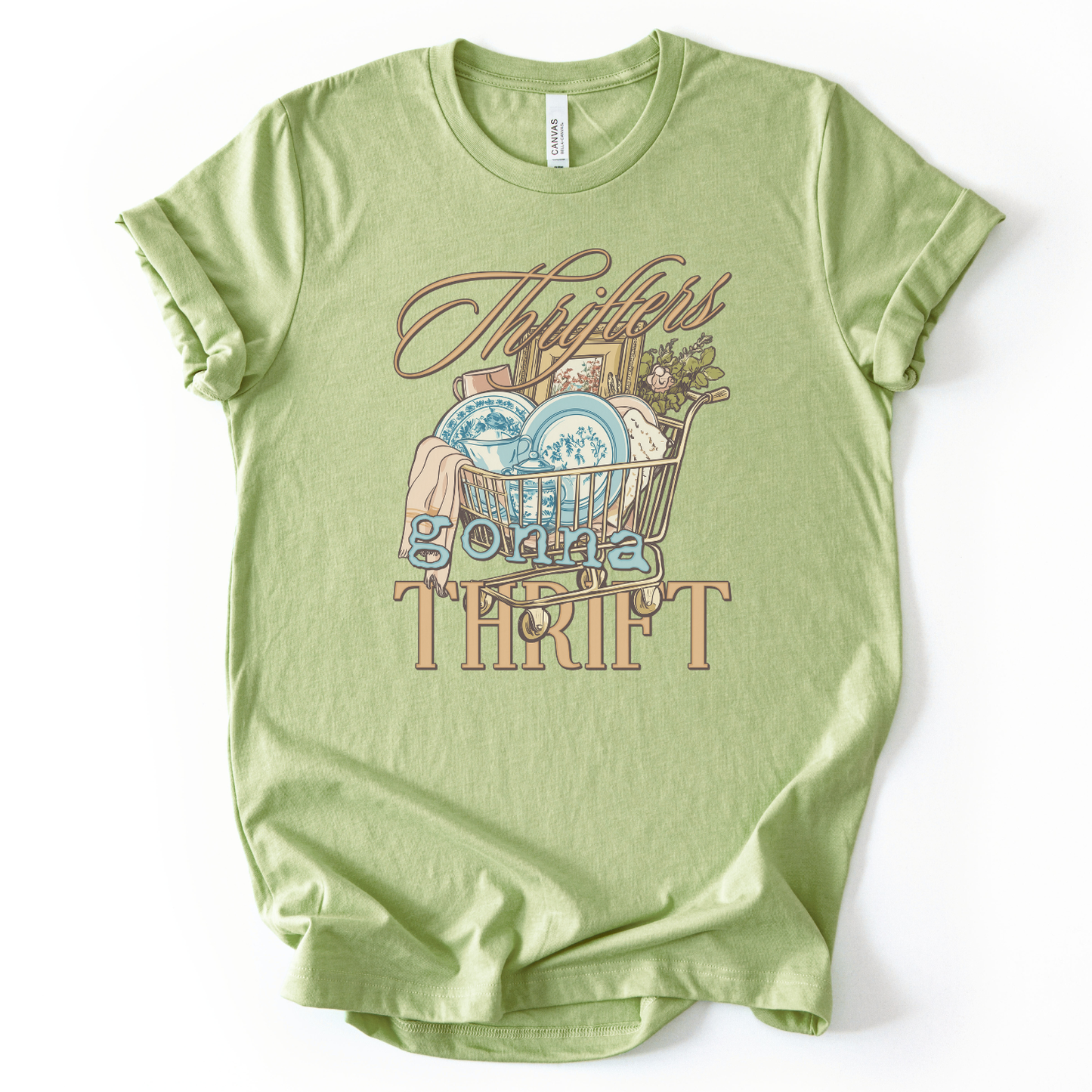 green t-shirt with graphic design and text on a white background.The trending graphic features an illustrated shopping cart filled with thrifted home decor finds, including blue toile-style plates, folded linens, framed artwork, and greenery. The phrase “Thrifters Gonna Thrift” appears in layered script and bold lettering, creating a warm, lifestyle-driven design that celebrates thrifting culture