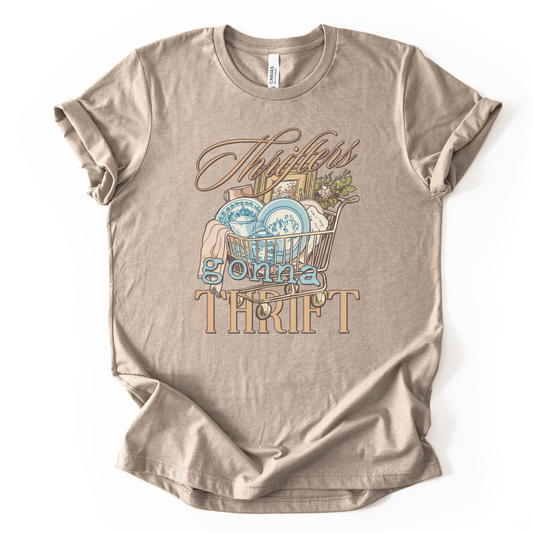 Beige t-shirt with graphic design and text on a white background.The trending graphic features an illustrated shopping cart filled with thrifted home decor finds, including blue toile-style plates, folded linens, framed artwork, and greenery. The phrase “Thrifters Gonna Thrift” appears in layered script and bold lettering, creating a warm, lifestyle-driven design that celebrates thrifting culture
