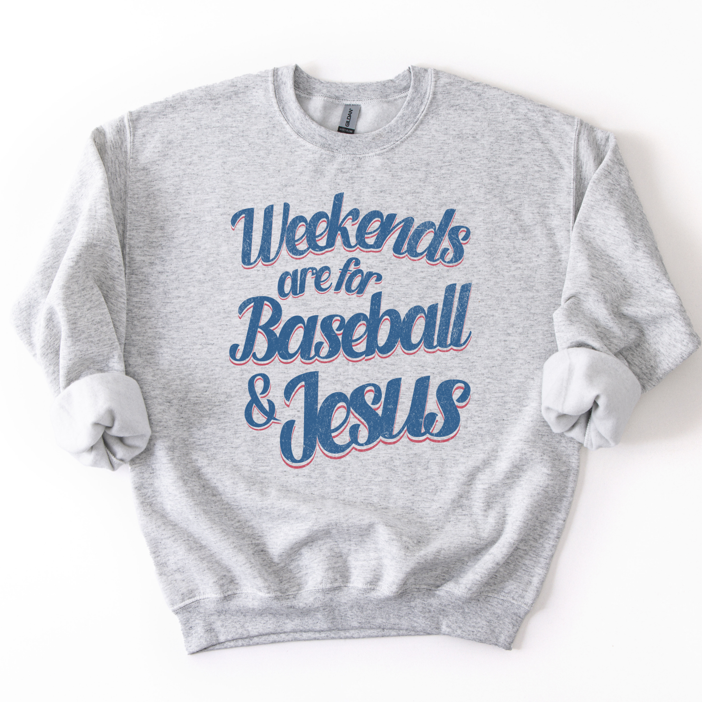 ash-colored crewneck sweatshirt with pink and blue retro script text reading “Weekends are for Baseball & Jesus,” styled flat with sleeves scrunched