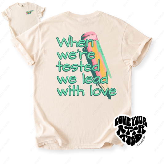 Painted pencil graphic with encouragement text “When We Are Tested We Lead With Love,” thick paint texture and high-contrast color, printed on a Comfort Colors® tee.