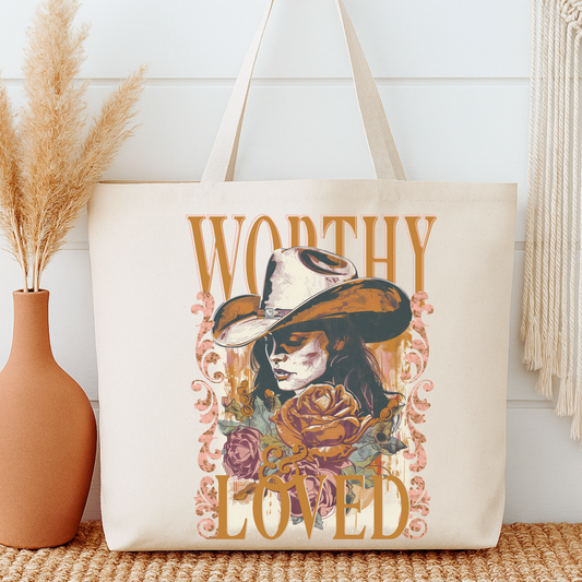 Tote bag with a design of a woman wearing a hat and flowers, with 'Worthy' and 'Loved' text, next to a vase with dried plants.
