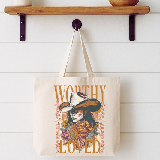 Tote bag with 'Worthy Loved' design hanging on a wooden shelf.