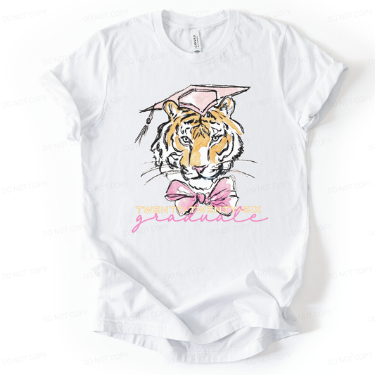 Graduation 2026 graphic t-shirt. The design features a detailed tiger illustration centered on the front, wearing a pink mortarboard cap with tassel and a large pink bow at the neck. Below the illustration, Twenty-Twenty-Six is printed in light lettering with a pink script graduate accent